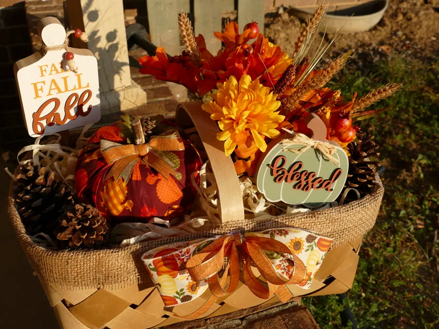 Harvest basket photographed from above, showing layered mums, wheat stems, a fabric pumpkin, and a sage 'blessed' sign.
