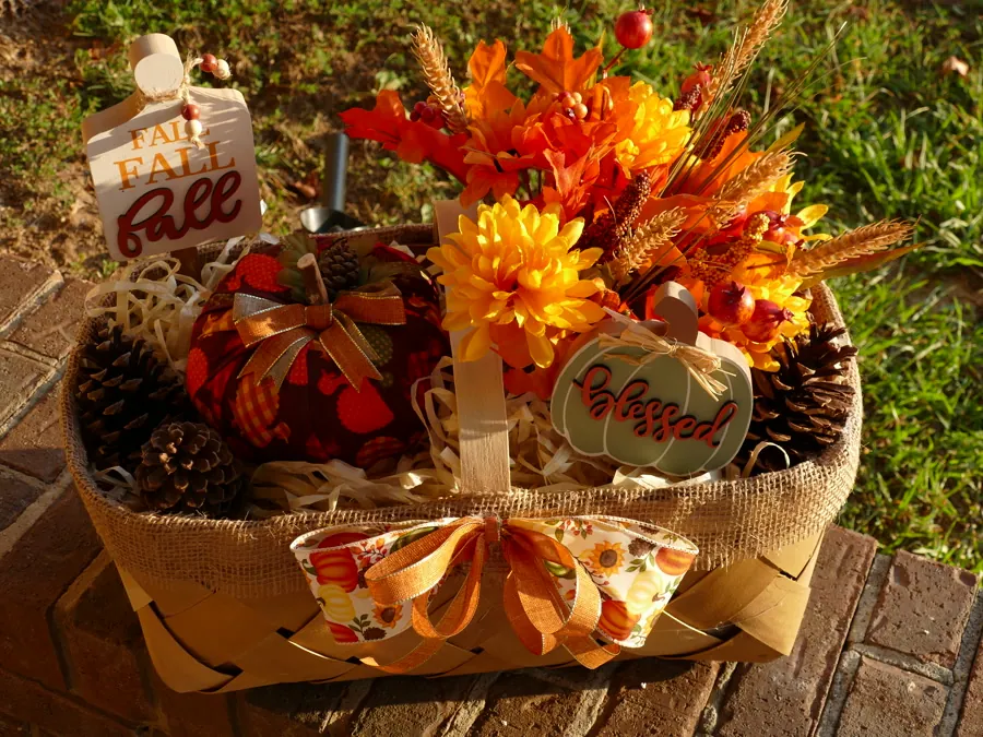 Woven harvest basket filled with a fabric pumpkin, pinecones, gold mums, and wheat, edged in burlap and set on a brick step in late afternoon light.
