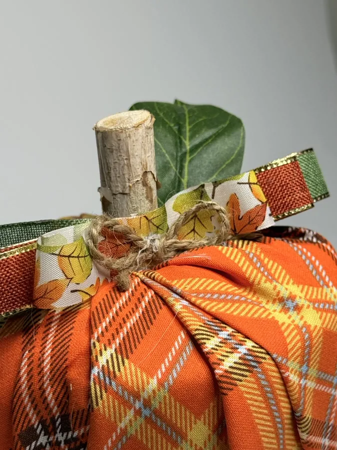 Close-up of a fabric pumpkin's birch stem, showing the jute twine wrap, a leaf-print ribbon, and a green silk leaf against orange plaid.