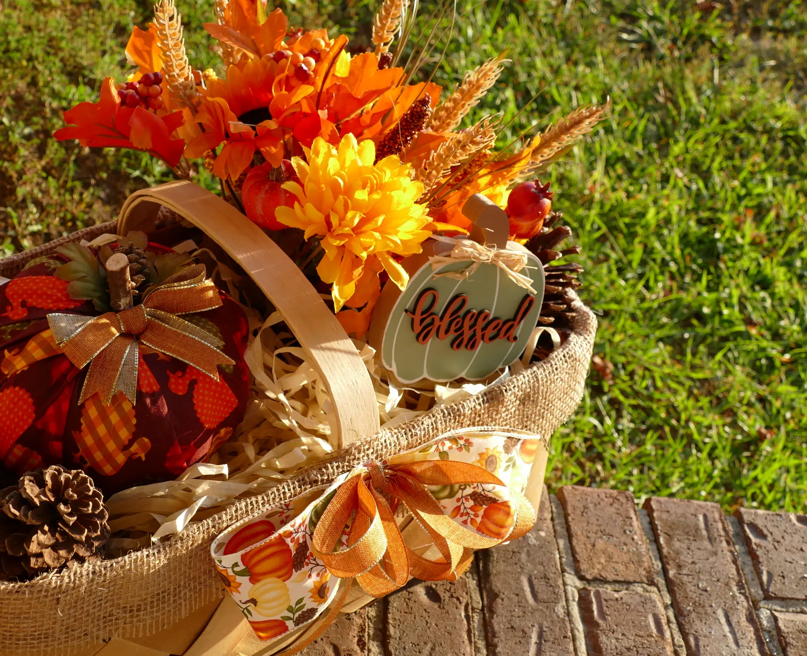 A handmade harvest basket wrapped in burlap, filled with a deep red fabric pumpkin, pinecones, gold and rust mums, wheat stems, and a sage green sign reading 'blessed', photographed outdoors in warm late-afternoon sunlight.