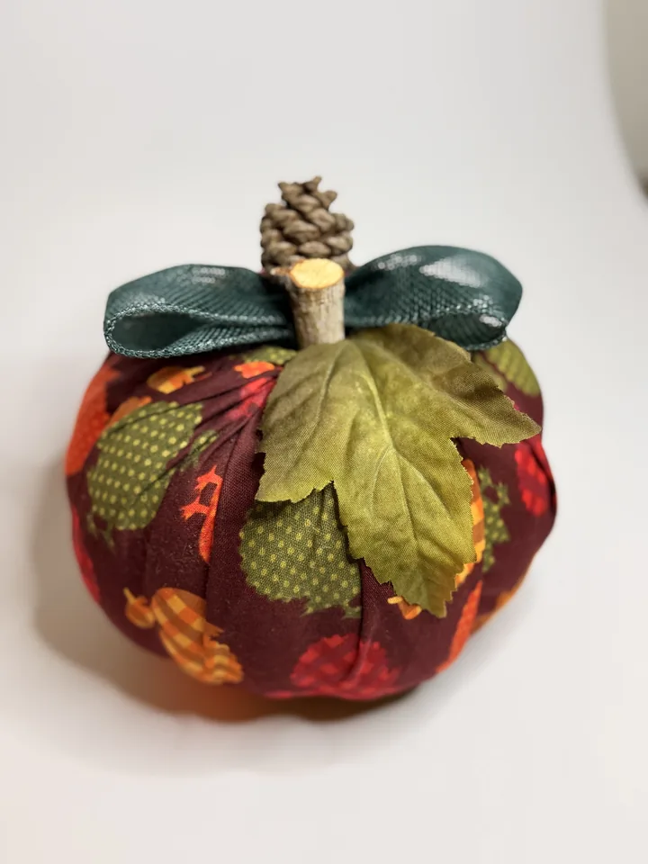 Burgundy fabric pumpkin printed with colorful patchwork acorns, topped with a teal bow, green silk leaf, and pinecone.