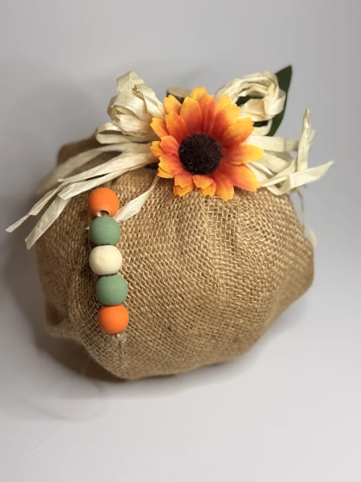 Natural burlap pumpkin with cream raffia, a burnt-orange sunflower, and orange, sage, and cream wood beads.
