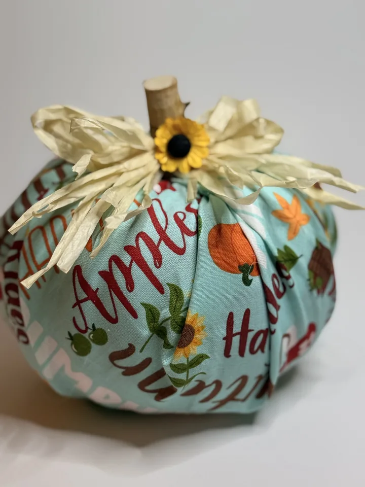 Aqua fabric pumpkin with harvest lettering, apples, pumpkins, and sunflowers, topped with cream raffia and a sunflower.