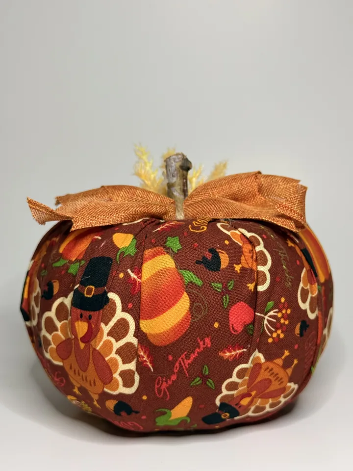 Brown fabric pumpkin printed with turkeys, pumpkins, and harvest treats, topped with a rust burlap bow and dried wheat.