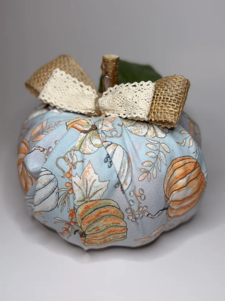 Powder-blue fabric pumpkin with watercolor pumpkins and leaves, topped with a burlap and ivory lace bow and a green ivy leaf.