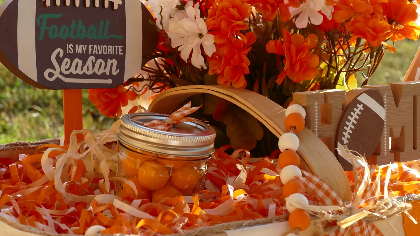 Close detail of a handmade arrangement: a ribbon-tied mason jar, painted wood bead garland, orange gingham bow, curled paper filler, and a woven basket rim lit by low afternoon sun.